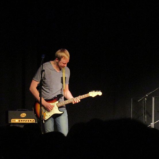 Ein Mann spielt eine E-Gitarre auf der Bühne bei schwachem Licht, mit einem Lautsprecher und Mikrofon in der Nähe. Das Publikum ist teilweise im Vordergrund zu sehen.