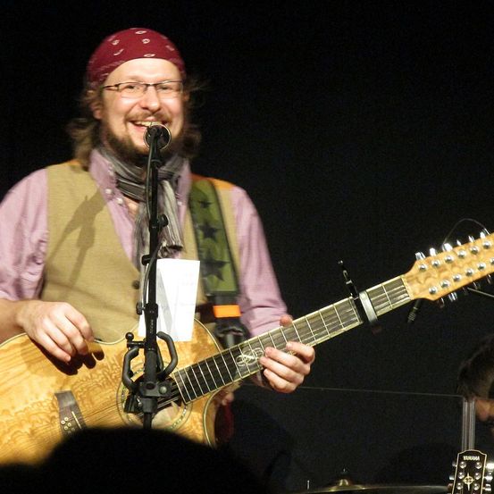 Ein Musiker spielt Gitarre und singt in ein Mikrofon auf der Bühne, trägt ein rotes Stirnband und eine Brille.