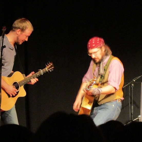 Zwei Männer spielen auf der Bühne mit Gitarren. Einer trägt ein graues T-Shirt, der andere ein rotes Stirnband und einen gelben Weste. Sie spielen vor einem dunklen Hintergrund und Mikrofonen.