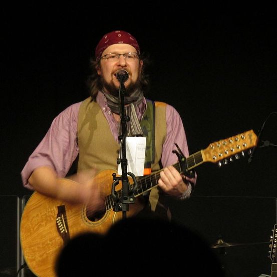 Ein Mann mit einem roten Bandana und Brille steht auf der Bühne und spielt Gitarre. Er singt in ein Mikrofon, während er die Gitarre mit beiden Händen hält.