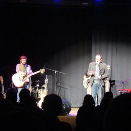 Ein Gitarrist und ein Sänger stehen mit einem Mikrofon auf der Bühne und treten vor einem Publikum auf. Der Gitarrist spielt eine akustische Gitarre, während der Sänger mit einem Mikrofon steht und ein Stück Papier in der Hand hält. Das Publikum sitzt vor der Bühne.