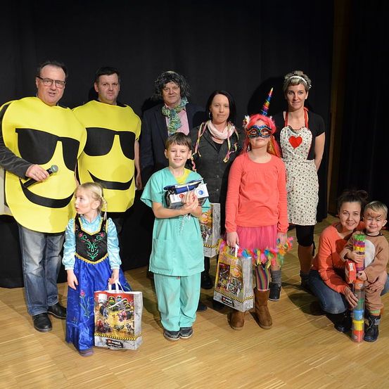 Eine Gruppe von Erwachsenen und Kindern in verschiedenen Kostümen steht auf einem Holzboden. Ein Mann in einem gelben Gesichtskostüm hält ein Mikrofon. Ein Junge hält eine Lego-Tasche. Ein Mädchen in einem roten Kleid trägt eine Maske. Ein anderes Mädchen trägt ein Einhornhorn.