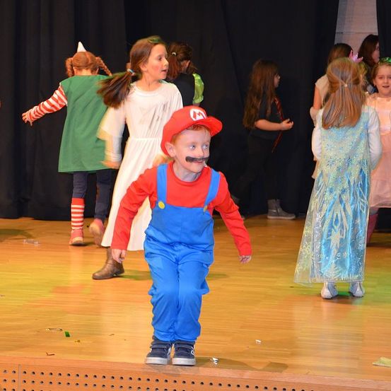Eine Gruppe von Kindern in verschiedenen Kostümen tritt auf der Bühne auf, darunter ein Junge in einem Mario-Kostüm mit einem falschen Schnurrbart.