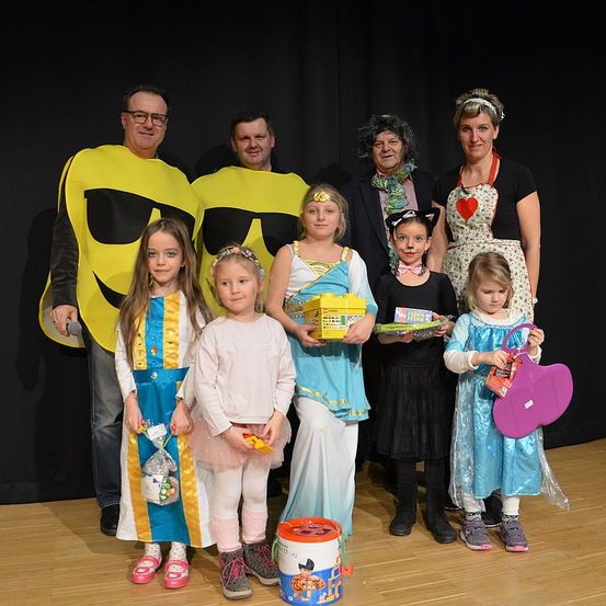 Eine Gruppe von Menschen in Kostümen posiert für ein Foto auf der Bühne. Zwei Erwachsene, die als Bananen verkleidet sind, stehen an den Seiten, während Kinder in verschiedenen Kostümen Geschenktüten und Spielzeug halten. Eine Frau in einer herzgedruckten Schürze steht hinter einem Eimer, und ein Mädchen in einem Katzenkostüm schaut zur Kamera.