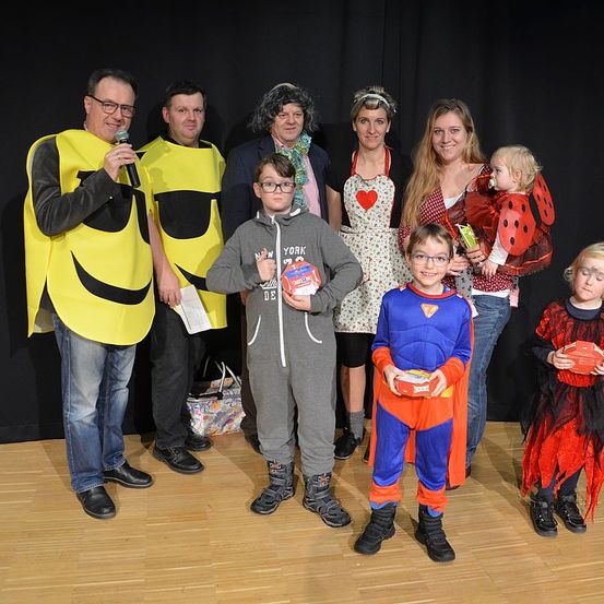 Eine Gruppe von Erwachsenen und Kindern in Kostümen steht zusammen auf einem Holzboden und hält einige Schachteln. Sie lächeln und posieren für ein Foto.
