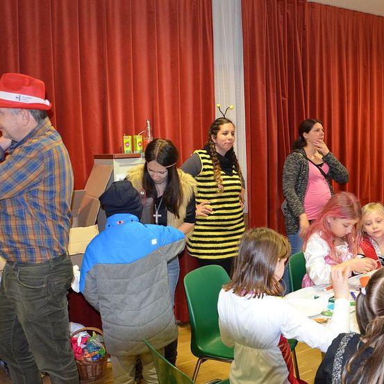 Eine Gruppe von Menschen, darunter ein Mann mit Hut, versammelt sich um einen Tisch, an dem Kinder in Stühlen sitzen. Ein Kind ist in einer Bienenkostüm verkleidet.