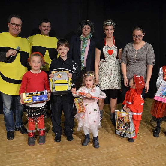 Eine Gruppe von Erwachsenen und Kindern in Kostümen posiert für ein Foto, einige halten Geschenke und Spielzeug. Die Erwachsenen tragen gelbe und schwarze Kostüme, während die Kinder verschiedene Kostüme tragen. Der Ort ist drinnen mit einem Holzboden und schwarzem Hintergrund.
