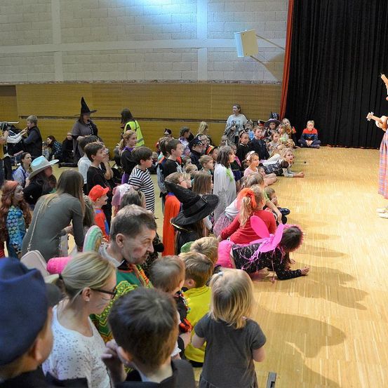 Eine Gruppe von Kindern und Erwachsenen versammelt sich in einem Saal für eine Aufführung. Die Kinder sitzen auf dem Boden und eine Frau steht auf der Bühne. Sie hält ein Musikinstrument. Das Publikum beobachtet sie. Der Saal hat Holzböden und Ziegelwände. Ein schwarzer Vorhang hängt an der Wand.