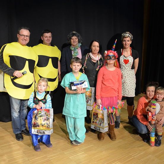 Eine Gruppe von Menschen in verschiedenen Kostümen, möglicherweise für ein Foto, darunter zwei Erwachsene in gelben Smiley-Kostümen, ein Kind in einer Arztuniform und andere in unterschiedlichen Outfits. Sie stehen und posieren auf einem Holzboden.