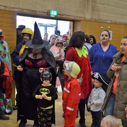 Mehrere Menschen sind in einem Raum versammelt. Eine Frau in einer Hexenmütze steht hinter einem Kind in einem Batman-Kostüm. Ein junger Junge in einem Feuerwehrmann-Anzug und ein Kind in einem Piratenhut sind vor ihnen. Der Raum hat einen Holzboden und eine gelbe Wand.