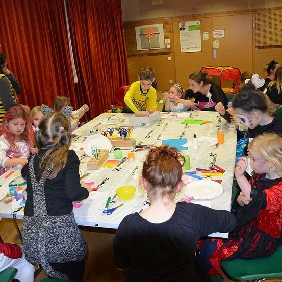 Eine Gruppe von Kindern und Erwachsenen versammelt sich um einen Tisch in einem Raum und beschäftigt sich mit einer Kunstaktivität. Der Tisch ist mit einer weißen Tischdecke bedeckt und hat verschiedene Kunstutensilien, einschließlich Scheren, Pinsel und Papier. Einige Kinder haben Gesichtsbemalung, und andere haben ihre Haare auf einzigartige Weise gestylt. Im Hintergrund befinden sich rote Vorhänge und eine Tafel mit angefügten Plakaten.