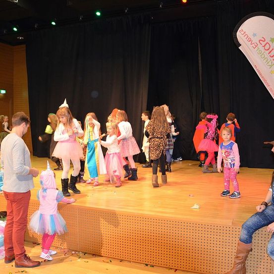 Mehrere Kinder in verschiedenen Kostümen stehen auf der Bühne, einige mit Requisiten. Ein Mann und ein Mädchen in Pink stehen in der Nähe. Die Bühne ist mit Konfetti geschmückt. Ein Banner befindet sich rechts.