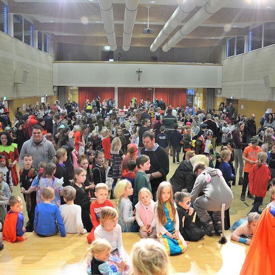 Ein großer Innenraum mit vielen Menschen in verschiedenen Kostümen, einschließlich Kindern, Erwachsenen und Polizeibeamten, versammelt für eine Veranstaltung. Einige sitzen auf dem Boden, andere stehen.