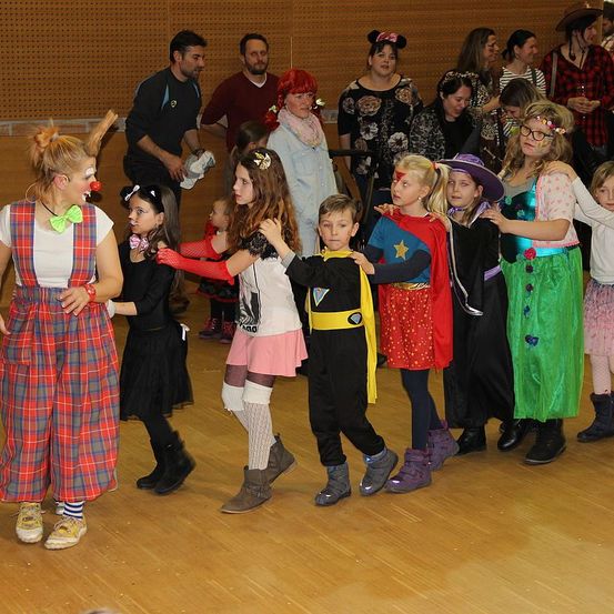 Eine Gruppe von Kindern und Erwachsenen in verschiedenen Kostümen steht in einer Reihe, wahrscheinlich für eine Halloween-Veranstaltung. Ein Clown und eine Frau in einem Kostüm sind vorne. Dahinter steht ein Mann in der Nähe der Wand.