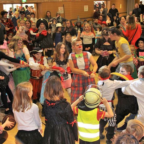 Eine große Gruppe von Kindern und Erwachsenen ist drinnen versammelt, möglicherweise bei einem Karneval oder einem thematischen Event. Viele sind in Kostümen gekleidet, darunter Clowns, Piraten und andere Charaktere. Eine Person in einem Clownskostüm mit roter Nase tanzt in der Mitte, während andere Händchen halten.