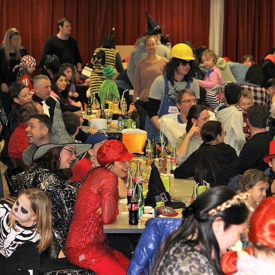 Ein Saal voller Menschen in Kostümen, die um Tische herumsitzen, einige lachen, ein Mann in einem gelben Hut und Sonnenbrille steht im Hintergrund, Flaschen und Becher stehen auf den Tischen.