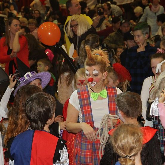 Ein Clown unterhält Kinder bei einer überfüllten Veranstaltung in Innenräumen, viele in Kostümen, während Erwachsene den Moment festhalten.