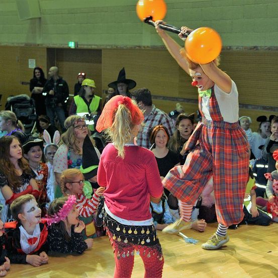 Ein Clown hält zwei orangefarbene Ballons und führt vor einer Menge von Kindern und Erwachsenen auf. Einige der Menge tragen Kostüme.