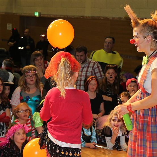 Ein Clown mit roter Nase und Mikrofon unterhält eine Menge, wobei ein Mädchen einen Ballon hält. Das Publikum, darunter Kinder, schaut aufmerksam zu.