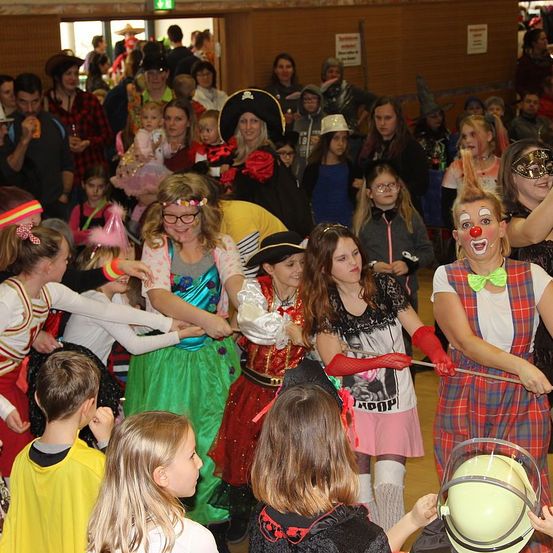 Eine Gruppe von Menschen ist in einem Saal versammelt, einige in Kostümen. Eine Frau in einer Piratenhaube hält ein Baby, während ein Clown mit roter Nase einen Stock hält. Eine weitere Frau in einem Kostüm und mit Brille lächelt. Eine Person in einem Helm ist ebenfalls anwesend.