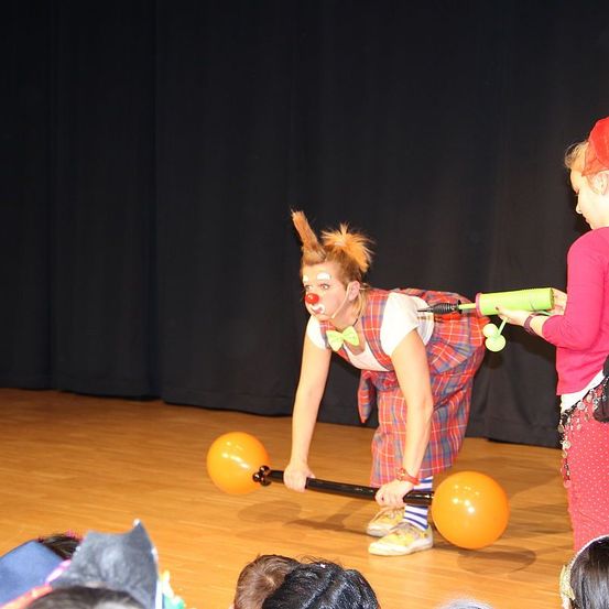 Ein Clown auf der Bühne mit einem großen orangefarbenen Ballon, einer roten Nase und einer Schleife. Ein Kind hält eine Ballonpistole neben ihnen. Das Publikum beobachtet von unten.
