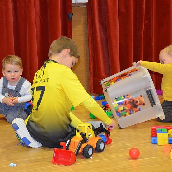 Drei kleine Kinder spielen mit Spielzeug auf einem Holzboden. Ein Kind hält einen Plastikbehälter mit Spielzeug, ein anderes ein Spielzeug-Lkw und ein drittes sitzt auf dem Boden.