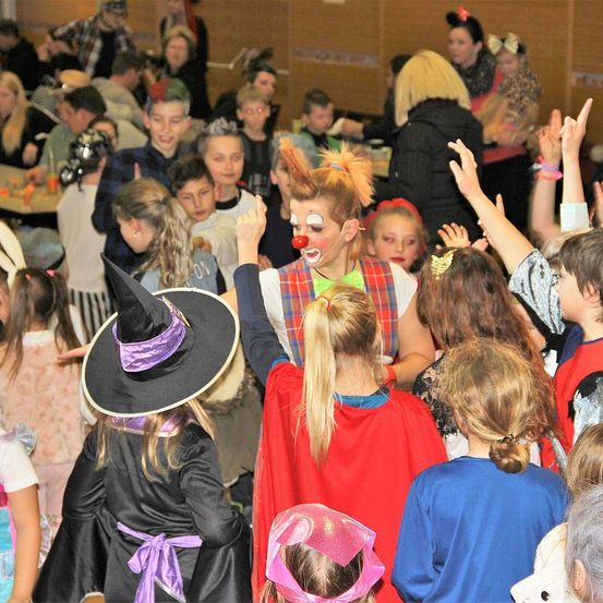 Eine Gruppe von Kindern und Erwachsenen versammelt sich in einem Raum, möglicherweise auf einer Party. Einige tragen Kostüme, darunter ein Clown und eine Hexe. Die Kinder heben ihre Hände und einige klatschen. Der Raum ist schwach beleuchtet.