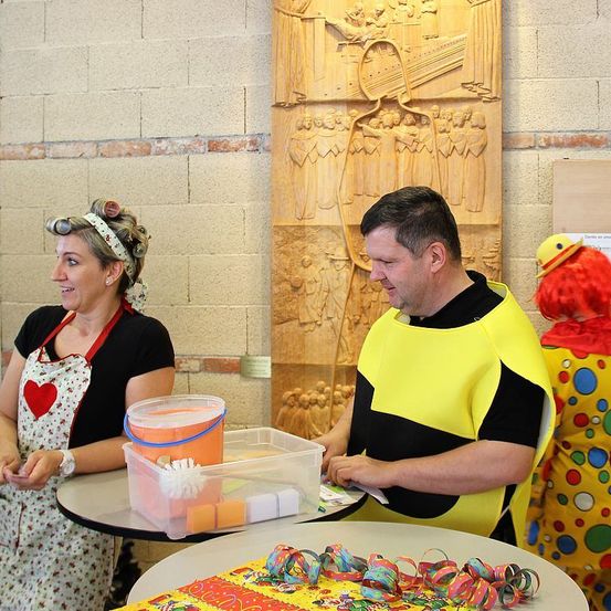 Ein Mann in einem gelben Clownskostüm spricht mit einer Frau in einer Blumenschürze. Ein Tisch mit Reinigungsutensilien steht zwischen ihnen. Ein Clownskostüm steht hinter ihnen. Eine Holzwand mit Schnitzereien befindet sich hinter dem Tisch.