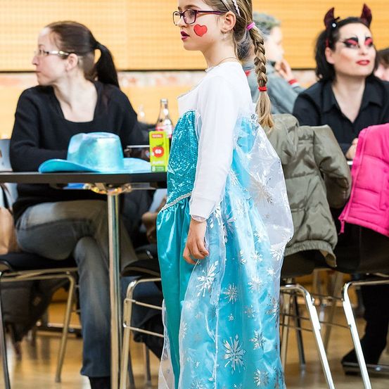 Ein kleines Mädchen in einem blauen Kleid steht in einem Raum mit Erwachsenen, einer trägt eine Teufelskostüm. Sie schaut sich um, während andere sitzen und reden. Ein Tisch hat einen Hut und eine Flasche.