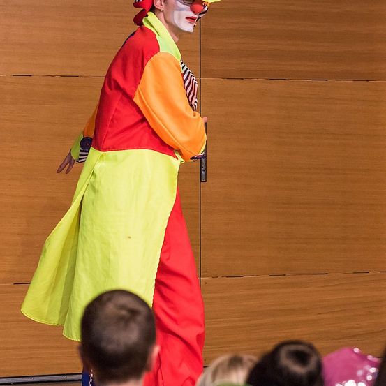 Ein Clown in einem bunten Kostüm tritt auf der Bühne vor einer Holzwand auf. Der Clown trägt einen Hut, eine Brille und einen gelben Mantel. Einige Kinder beobachten die Vorstellung.