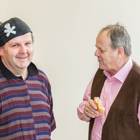 Zwei Männer stehen vor einer weißen Wand. Der Mann auf der linken Seite trägt einen schwarzen Piratenhut mit einem weißen Totenkopf und gekreuzten Knochen. Der Mann auf der rechten Seite hält ein Sandwich und trägt ein rosa Hemd und eine braune Weste.