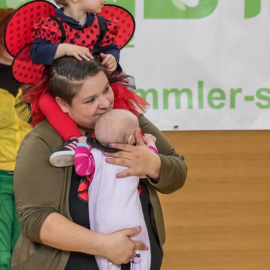 Eine Frau hält ein Baby, während ein weiteres Kind in einem roten Kostüm auf ihrem Kopf sitzt. Ein Banner ist im Hintergrund sichtbar.