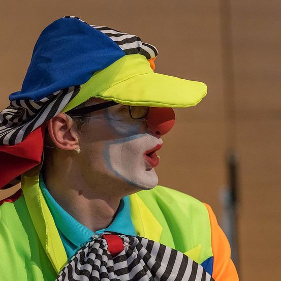 Ein Clown mit einem mehrfarbigen Hut, weißem Gesichtsmal und einem neongrünen Weste mit Fliege steht in einem Raum.