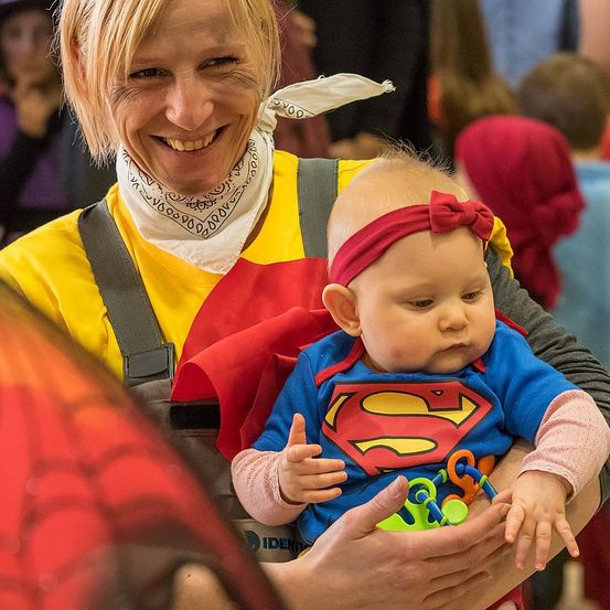 Eine lächelnde Frau, die eine gelbe Schürze mit einem Superman-Logo trägt, hält ein als Superman verkleidetes Baby. Das Baby hat eine rote Schleife auf dem Kopf und hält ein Spielzeug. Der Hintergrund ist unscharf.