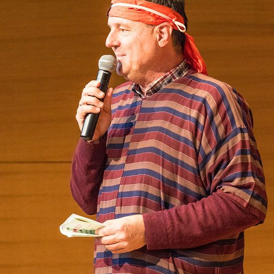 Ein Mann mit rotem Stirnband spricht in ein Mikrofon und hält ein Stück Papier in der Hand.