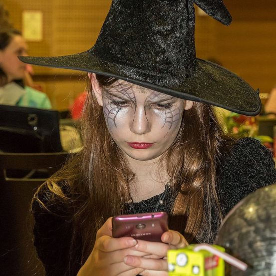 Ein Mädchen in einem Hexenkostüm schaut auf ihr Handy, mit Spinnennetzen auf dem Gesicht gemalt. Im Hintergrund sitzen andere Personen am Tisch.