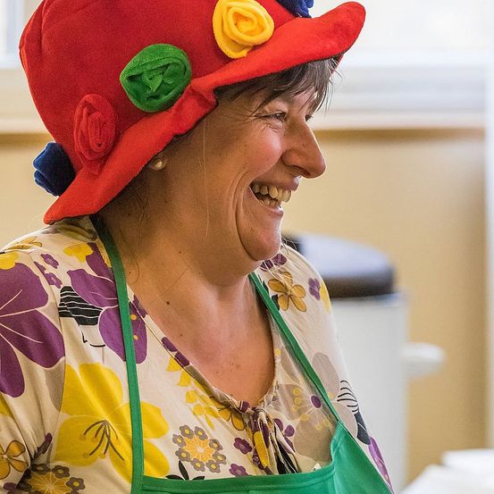Eine Frau trägt einen roten Hut mit bunten Blumen, ein Blumenhemd und eine grüne Schürze. Sie lächelt und scheint sich in einem Raum zu befinden.