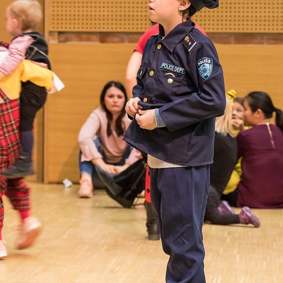 Ein junger Junge in einer Polizeiuniform steht auf einer Bühne mit anderen Kindern. Hinter ihm sitzt eine Frau auf dem Boden. Im Hintergrund läuft ein Kleinkind vorbei.