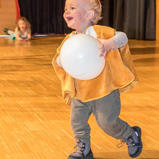 Ein junger Junge in einem gelben Westchen hält einen weißen Ballon und geht auf einem Holzboden. Ein Mädchen sitzt im Hintergrund.