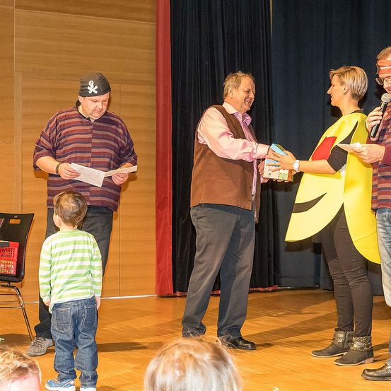 Ein Mann in einer Piratenmütze liest von einem Papier, während ein anderer Mann einer Frau in einer Bananenkostüm ein Geschenk überreicht. Ein Mann hält ein Mikrofon, um zu sprechen.