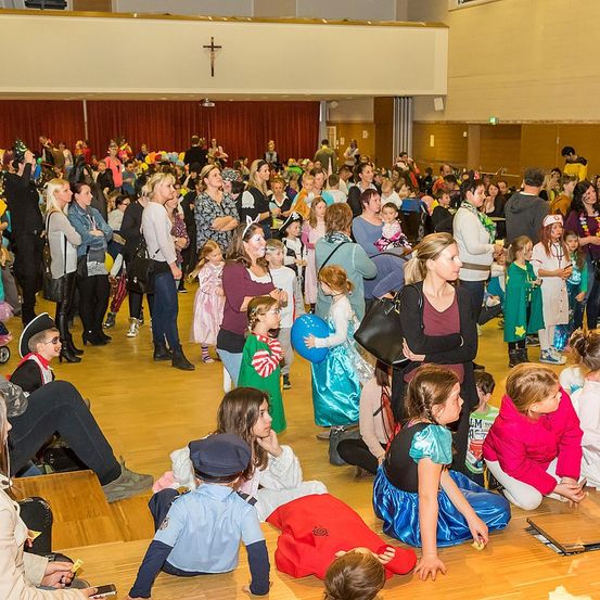 Eine große Menschenmenge, darunter Kinder in Kostümen, versammelt sich in einem Saal, einige sitzen auf dem Boden und andere stehen, was eine festliche Atmosphäre schafft.