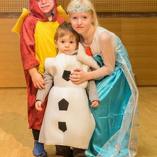Drei Kinder in Kostümen posieren für ein Foto. Ein junger Junge in einem Drachenkostüm steht neben einem Kleinkind, das als Olaf gekleidet ist. Ein als Elsa gekleidetes Mädchen hält das Kleinkind. Sie sind drinnen auf einem Holzboden.