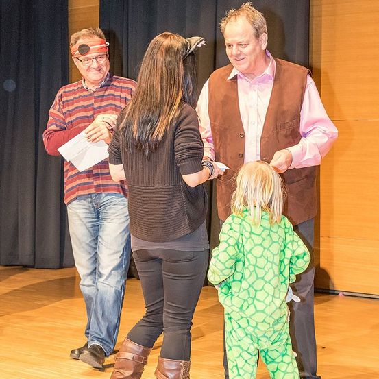 Drei Personen auf einer Bühne, einer davon ist ein Mann in einem braunen Vest, eine andere ist eine Frau in Schwarz und ein Kind in Grün. Der Mann hält ein Stück Papier.