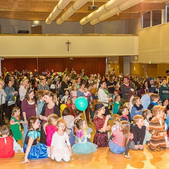 Eine große Gruppe von Menschen, meist Kinder in Kostümen, versammelt sich drinnen. Einige stehen und andere sitzen auf dem Boden. Eine Frau hält einen grünen Ballon, und ein Kreuz ist an der Wand montiert.