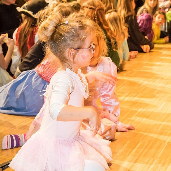 Ein kleines Mädchen in einem rosa Kleid und mit Brille sitzt auf einem Holzboden und sieht sich eine Aufführung mit anderen Kindern an. Einige sind in Kostümen und der Hintergrund ist unscharf.