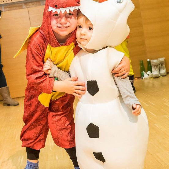 Ein Junge, verkleidet als Drache, hält einen Kleinkind in einem Schneemannkostüm in einem Raum.