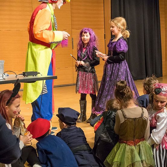 Ein Clown in bunter Kleidung unterhält Kinder in Kostümen auf einer Bühne mit dunklem Vorhang als Hintergrund.