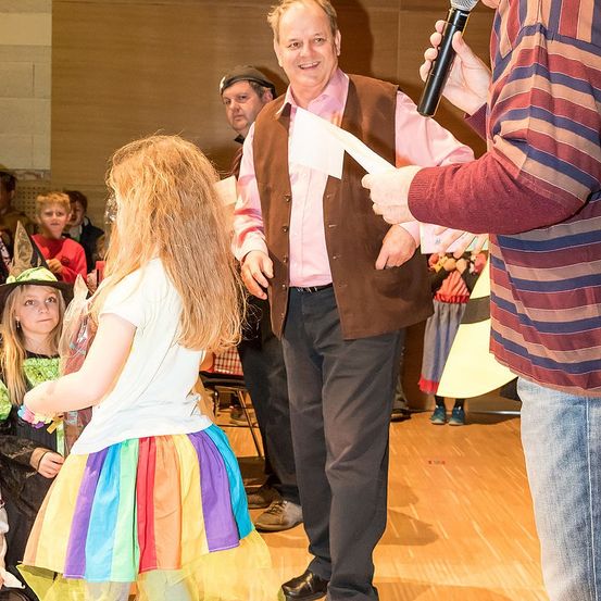 Ein Mann gibt einem Mädchen in einem Regenbogen-Tutu ein Papier, während ein anderer Mann ein Mikrofon hält. Andere Menschen sind in einem Saal anwesend, einige sind in Kostümen gekleidet.