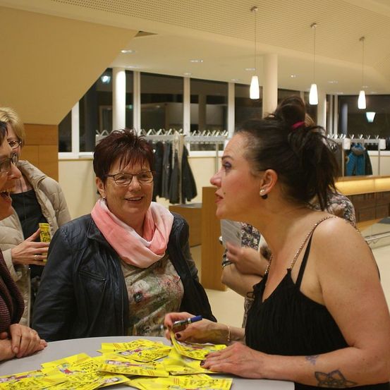 Fünf Frauen versammeln sich in einem Einzelhandelsgeschäft um einen Tisch. Eine Frau hält Stift und Papier, eine andere Frau hat ein gelbes Ticket. Im Hintergrund sind Kleiderständer und Lampen zu sehen.