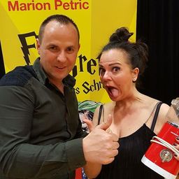 Ein Mann und eine Frau posieren für ein Foto vor einem Schild, das Marion Petric liest. Der Mann zeigt Daumen hoch und die Frau hält ein rotes Gerät.